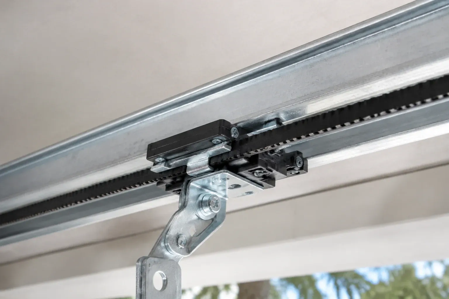 Black plastic trolley and bent galvanized bracket on a steel opener rail with a toothed black belt, against a ceiling. — Belt-Drive Opener Repair, Oakville